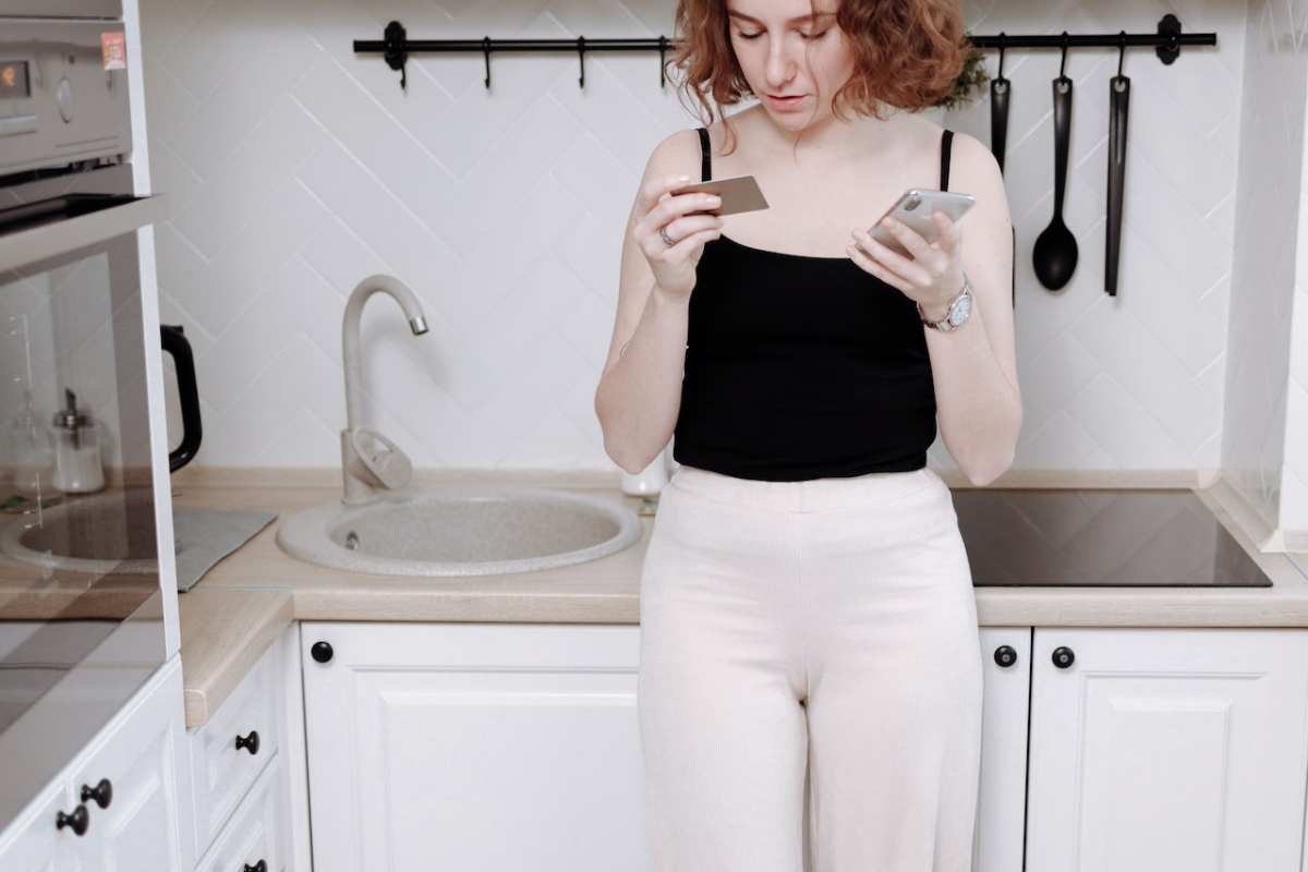 httpswww.pexels.comphotowoman standing in the kitchen while holding cellphone and a card 6237996